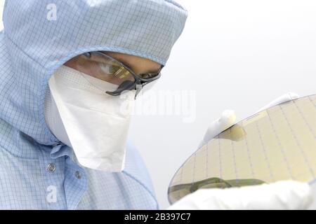 An engineer working in a clear room wearing a special uniform and protective glasses holds silicon wafer with microchips in hands in gloves Stock Photo