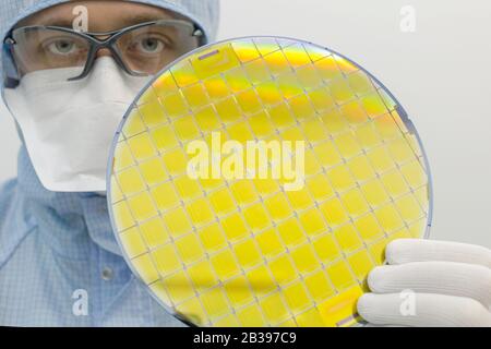 An engineer working in a clear room wearing a special uniform and protective glasses holds silicon wafer with microchips in hand in glove Stock Photo