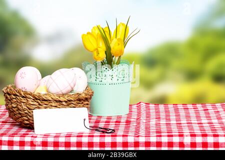 Easter card template. Colorful easter eggs in a wicker basket, yellow tulips and a empty tag on a red checkered tablecloth against abstract natural sp Stock Photo