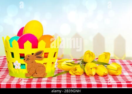 Happy Easter greeting card template. Colorful easter eggs in a basket with a bouquet of yellow tulips on picnic tablecloth over abstract blurred sprin Stock Photo