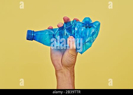 Hand squeezing blue plastic bottle on yellow background. Zero waste and consumption reduction of plastic concept. minimal, copy space Stock Photo