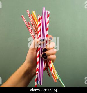 A handful of plastic disposable drinking straws in a female hand on a green background. Not green things, environmental pollution concept. Stock Photo