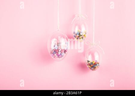 Easter eggs are transparent and filled with small colorful flowers hanging on a rope on a pink background. Minimalistic holiday decoration. Stock Photo