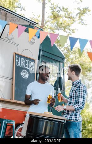 cheerful multicultural friends holding bottles of beer near french fries and food truck Stock Photo