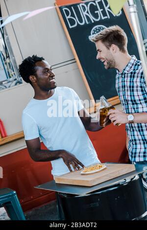 positive multicultural men toasting bottles of beer near french fries and food truck Stock Photo