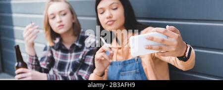 panoramic shot of pretty friends smoking cigarettes and taking selfie Stock Photo