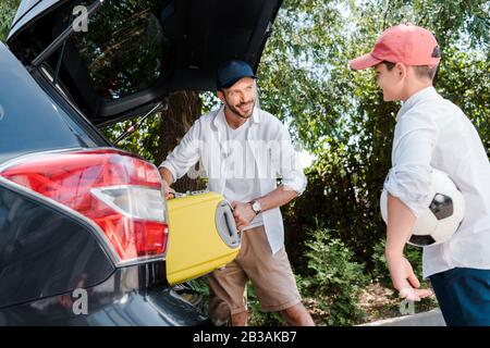 selective focus of happy man in cap putting travel bag in car trunk near son Stock Photo