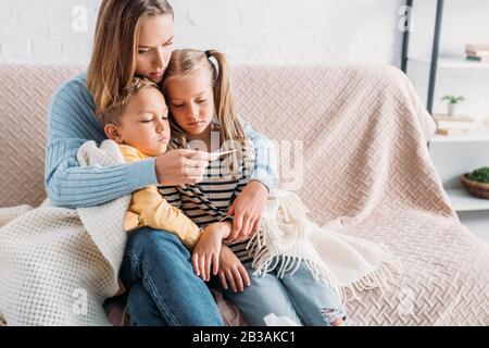 upset woman looking at thermometer while sitting on sofa with diseased children Stock Photo