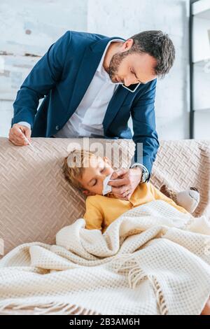 caring father wiping nose of sick son while holding thermometer and talking on smartphone Stock Photo