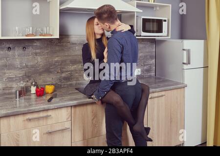 Young couple in love kisses in the kitchen. A slender white woman sits on a table and hugs with her legs a Caucasian man who is standing opposite her. Stock Photo