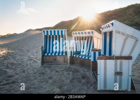 Beach chairs at sunrise on Sylt island, Germany. Wicker chairs on an empty beach at the North Sea and sun rays. Stock Photo