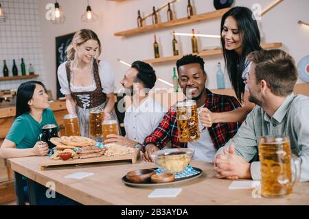 two attractive waitresses serving beer for multicultural friends in pub Stock Photo