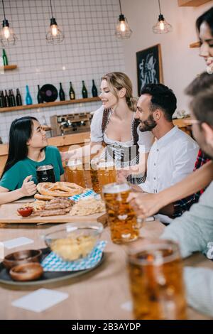 two waitresses in traditional german costume serving beer for multicultural friends in pub Stock Photo