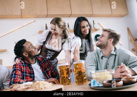 two cheerful waitresses in traditional german costumes serving beer for multicultural friends in pub Stock Photo