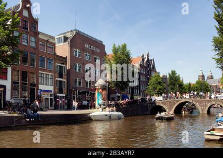 View of people riding a boat on canal doing a cruise tour in Amsterdam. Many people hang out on street and crossing Armbrug bridge. It is a sunny summ Stock Photo