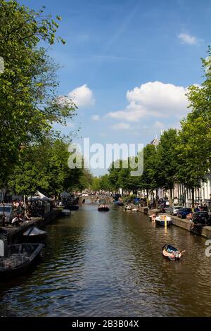 View of people riding small boats on canal doing a cruise tour in Amsterdam. Cloudy blue sky and trees are also in the view. It is a sunny summer day. Stock Photo
