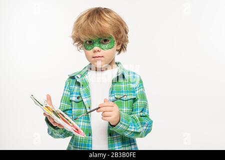 adorable boy with gecko mask painted on face holding palette and paintbrush while looking at camera isolated on white Stock Photo