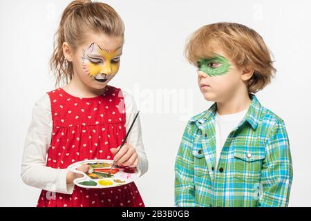 adorable kid with cat muzzle face painting holding palette and paintbrush near boy with painted gecko mask isolated on white Stock Photo