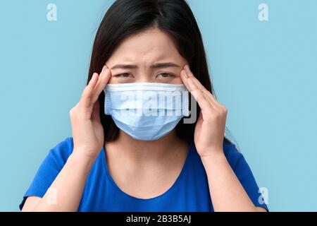 Coronavirus Concept. Chinese woman wearing medical mask standing isolated on grey having headache close-up Stock Photo