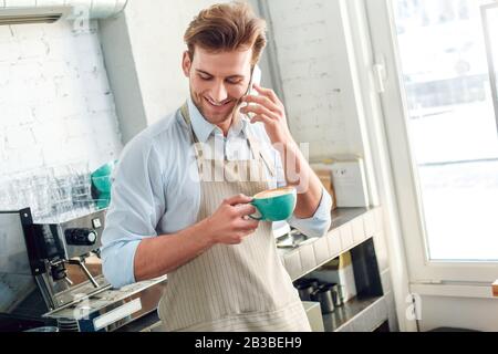 Coffee Passion. Young barista at coffee shop with cup of hot coffee talking on smartphone cheerful Stock Photo