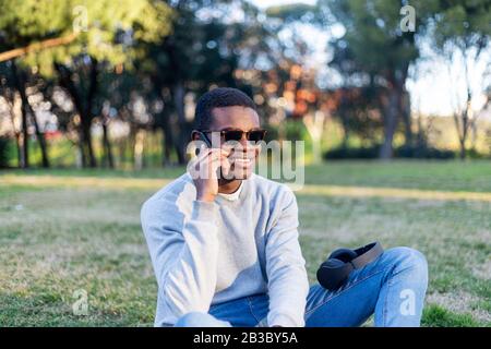 Fashionable male with dark skin and sunglasses sitting outdoors checking his news feed or messaging via social networks using cell phone Stock Photo