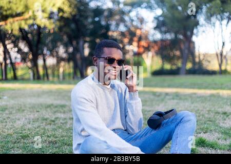 Fashionable male with dark skin and sunglasses sitting outdoors checking his news feed or messaging via social networks using cell phone Stock Photo