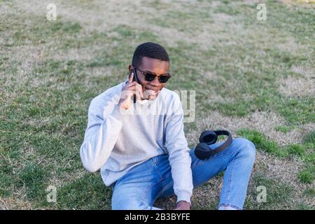 Fashionable male with dark skin and sunglasses sitting outdoors checking his news feed or messaging via social networks using cell phone Stock Photo