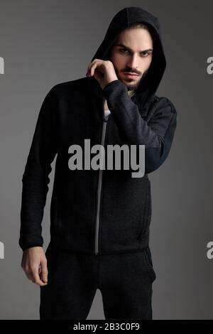 beautiful casual man wearing tracksuit standing with his hood on head and touching his chin on gray studio background Stock Photo