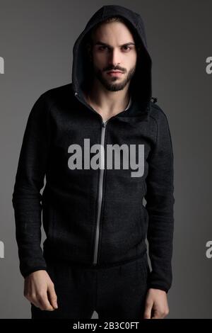 attractive casual man wearing tracksuit standing and staring at camera with his hood on head on gray studio background Stock Photo