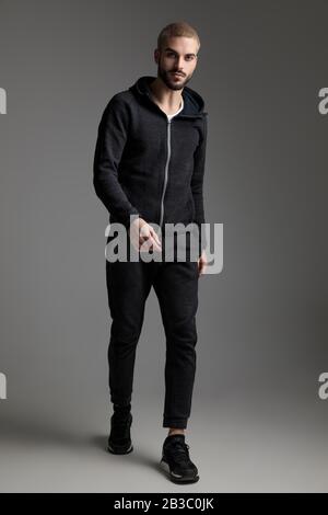 attractive casual man wearing tracksuit walking and staring at camera on gray studio background Stock Photo