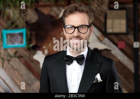 Cheerful groom laughing while wearing tuxedo and glasses, standing on coffeeshop background Stock Photo