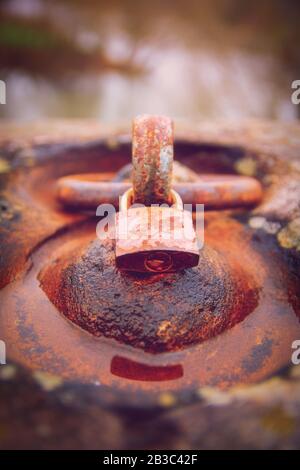 Close up of a rusty padlock locked around an aged rusty iron ring in a puddle of water. Time and aging concept. Stock Photo