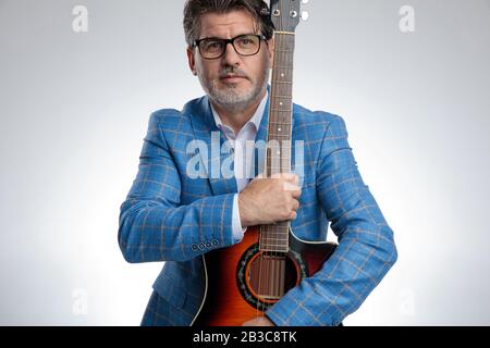 portrait of a charming formal business man wearing a light blue suit and eyeglasses standing and holding his guitar tight while looking at camera seri Stock Photo