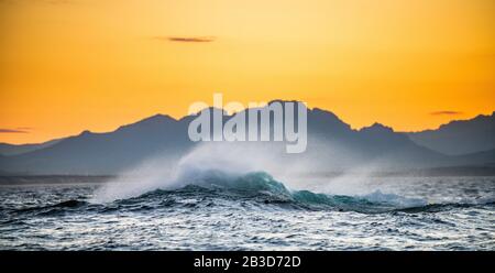 Seascape.  Red dawn sky, waves crashing with splashes against stones, silhouettes of mountains on the horizon.  South Africa.