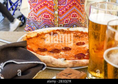 A woman sits at an outdoor cafe table with pepperoni pizza and beer on the table in front of her. Stock Photo