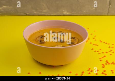 Pea soup with mushrooms in a pink plate stands on a yellow background, around the scattering of lentil seeds Stock Photo