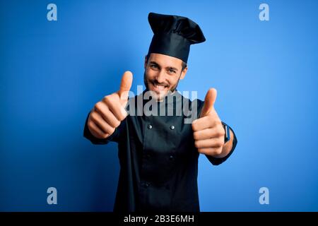 Young handsome chef man with beard wearing cooker uniform and hat over blue background approving doing positive gesture with hand, thumbs up smiling a Stock Photo