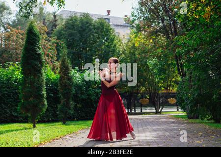 Woman ballerina in red ballet dress dancing in pointe shoes in autumn park. Stock Photo