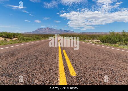 Long straight road in the desert, leading to a beautiful mountain range under a blue sky with clouds Stock Photo