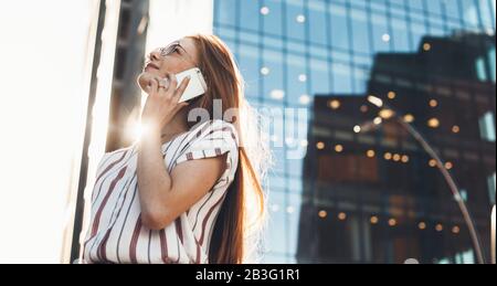 Caucasian red haired businesswoman with freckles is speaking on phone while looking away through eyeglasses Stock Photo