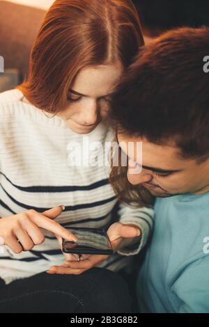 Red haired caucasian girl with freckles embraced by her lover is looking for something in her mobile phone Stock Photo