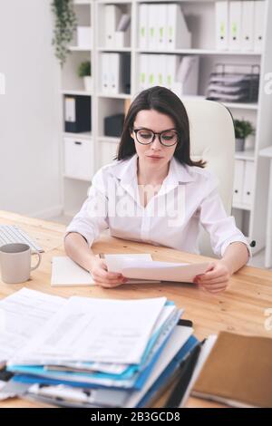 Concentrated young businesswoman in eyeglasses sitting at desk in office and analyzing contact content Stock Photo