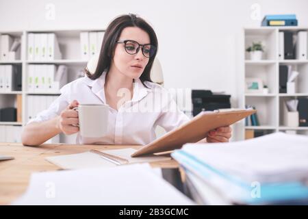 Busy young office woman sitting at table and reading papers while drinking coffee in office Stock Photo