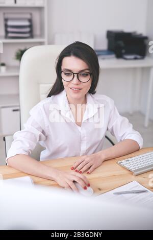 Smiling young smart lady in glasses sitting at desk and using modern computer in office Stock Photo