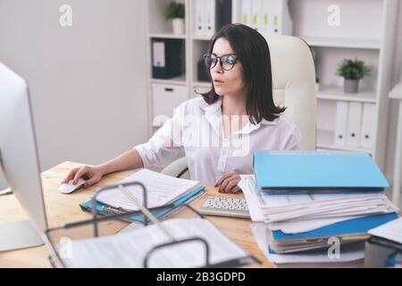 Busy young woman in blouse sitting at desk and using computer while working on pile of documents Stock Photo