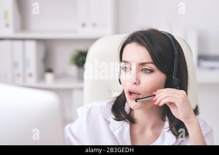 Young call center operator in microphone headset answering online call and checking information on computer monitor Stock Photo