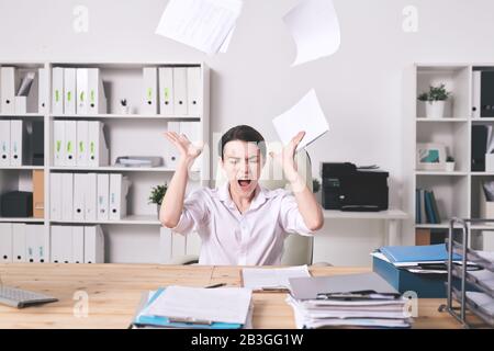 Frustrated young female manager annoyed with paperwork throwing papers and screaming with closed eyes in office Stock Photo