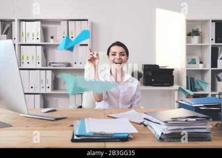 Cheerful excited young businesswoman sitting at desk full of documents and throwing paper plane in office Stock Photo