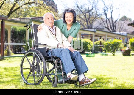 Asian and african nurses helping elder man on wheelchair at rehab facility garden. Stock Photo