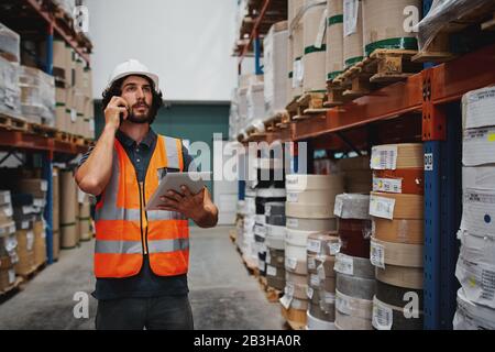 Warehouse manager tracking supply order details using digital tablet while in conversation over mobile phone in warehouse Stock Photo